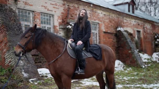 フード付きの黒いガウンを着たカリスマ的髭男が馬に乗って座っている — ストック動画