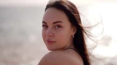 Güzel, tombul, uzun siyah saçlı bir kadın denizin kenarında kameraya bakıyor.