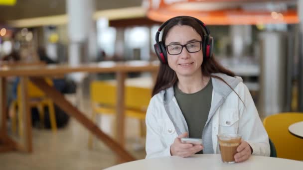 Happy lady is resting in cafe, drinking coffee and listening to her favorite music by headphones — Stock Video