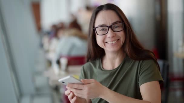 Θετική γοητευτική γυναίκα χαμογελά στην κάμερα και διορθώνει τα γυαλιά της, κρατώντας το smartphone στο χέρι — Αρχείο Βίντεο