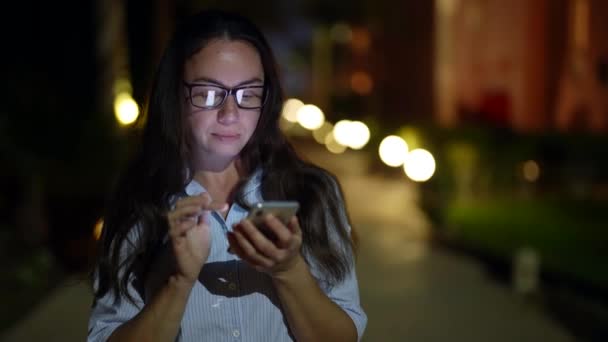 Wanita dewasa mengirimkan pesan dalam percakapan online oleh smartphone di jalan di malam hari — Stok Video