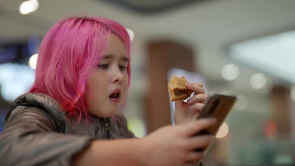 Close-up van een tienermeisje met roze haar dat goedkoop eten eet in een winkelcentrum en sociale netwerken bekijkt op haar telefoon. Internetafhankelijke generatie — Stockvideo