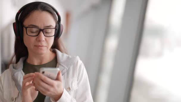 Mujer adulta con gafas y auriculares está escuchando música por teléfono inteligente y bailando — Vídeo de stock