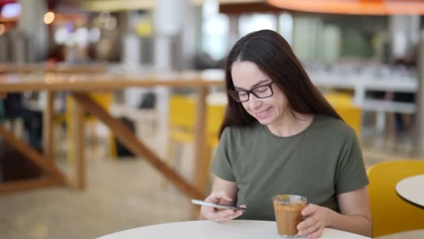 Gündüz vakti gözlüklü ve akıllı telefonlu modern bir kadın kafede dinleniyor ve internette sohbet ediyor. — Stok video