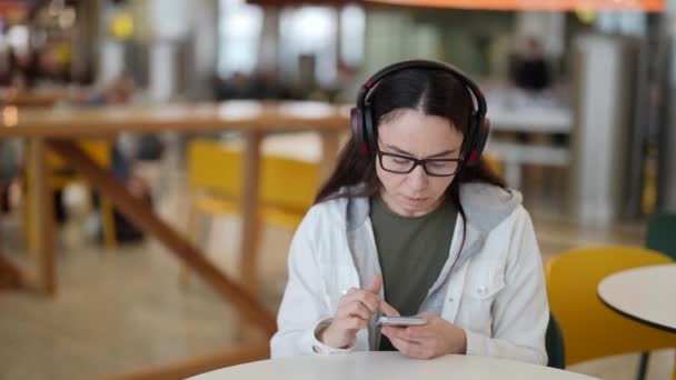 Взрослая женщина сидит за столом в кафе и слушает музыку по беспроводному наушнику — стоковое видео