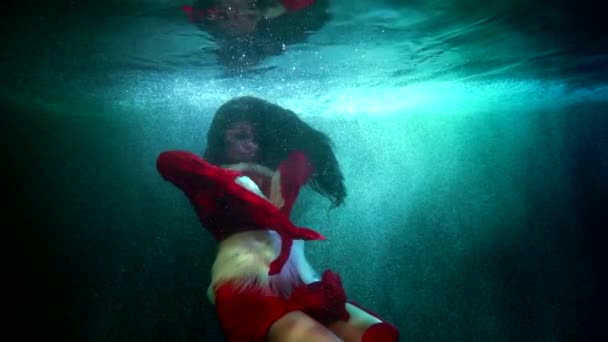 Hermosa mujer morena en vestido rojo se mueve con gracia en la piscina o el lago, tiro bajo el agua — Vídeo de stock