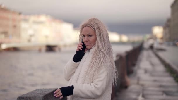 Seriöse blonde Frau mit langen weißen Haaren telefoniert im Winter am Stadtkai — Stockvideo