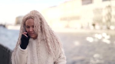 Yetişkin sarışın kadın, şehir caddesindeki nehir kenarında cep telefonundan arıyor.