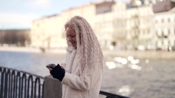 Jovem mulher loira está usando smartphone no dique da cidade no dia de inverno — Vídeo de Stock