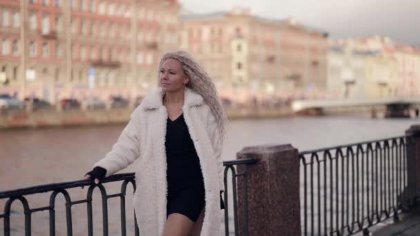Wanita pirang seksi bermantel bulu putih berdiri di tanggul sungai kota di hari yang cerah, wanita trendi — Stok Video