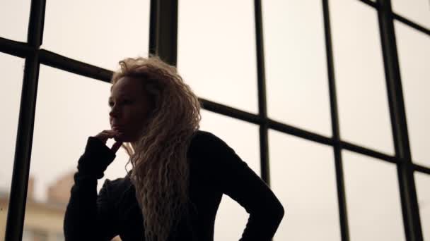 Стомлена блондинка з довгим волоссям, портрет на фоні великого вікна — стокове відео