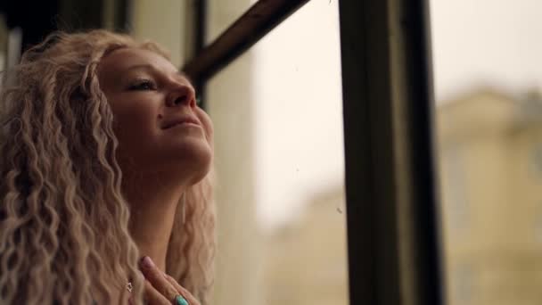 Uzun sarı saçlı güzel moda kadın pencereye bakıyor ve gülümsüyor. — Stok video