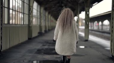 Beyaz kürklü yalnız bir kadın boş eski tren istasyonunda yürüyor.