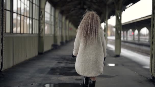 Lonely woman in white fur coat is walking in empty old railway station, urbanistic loft location — Stock Video