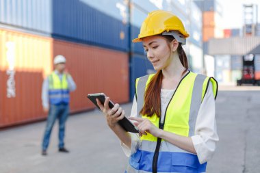 Women worker use tablet in uniform wearing safety helmet using laptop checking containers loading. Area logistics import export and shipping.