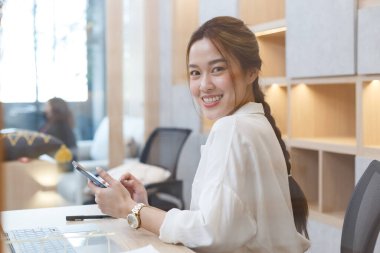 Genç Asyalı gülümseyen iş kadını ofis masasında akıllı telefon kullanıyor. Başarılı kız cep telefonundan mesaj okuyor. Boşluğu kopyala.