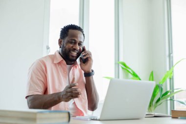 Siyahi Afrika kökenli Amerikalı cep telefonu alıyor ve ofis masasında dizüstü bilgisayarla araştırma yapıyor..