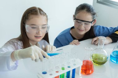 Çocuk bilim öğrencileri, beyaz laboratuvar odasında sıvı kimyasal deneyler üzerinde çalışıyorlar. Öğrenci kız elinde biyo-teknoloji ile test tüpü tutuyor. Bilim eğitimi kavramı.