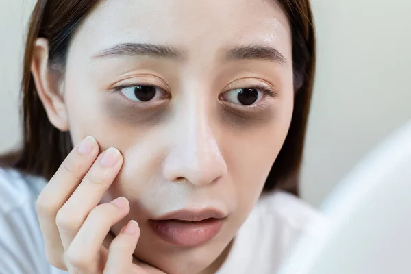 Close up face of asian young female stress looking mirror check have dark eye circles on face from staying up sleepy late. from hard work, no time to sleep.