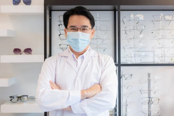 Portrait of asian man professional optician selling wear spectacles and protective face mask crossed arm standing at optical shop.