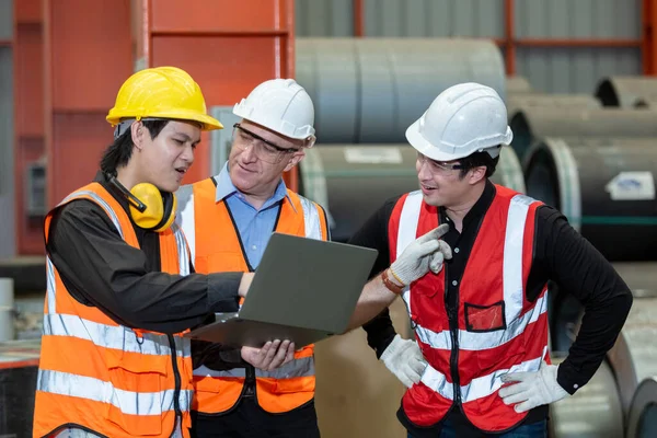 Three man industrial engineers wear hard hats and uniform using laptop talking project of factory inside heavy industry manufacturing. Supervisor teaching work to employee.