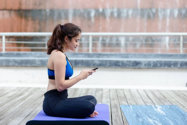Young sport woman sportswear using smartphone sitting on yoga mat outdoor in garden. relax after yoga and exercise.