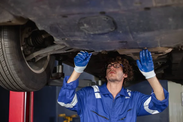 young man auto mechanic repairman wearing eyeglasses in uniform checking car suspension repair in auto garage, service and maintenance concept.