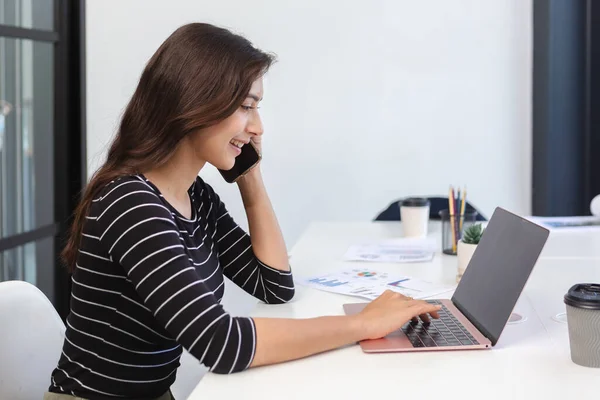 Smiling asian business woman talk on cell phone and using laptop. female freelance professional calling with customer work inquiries.