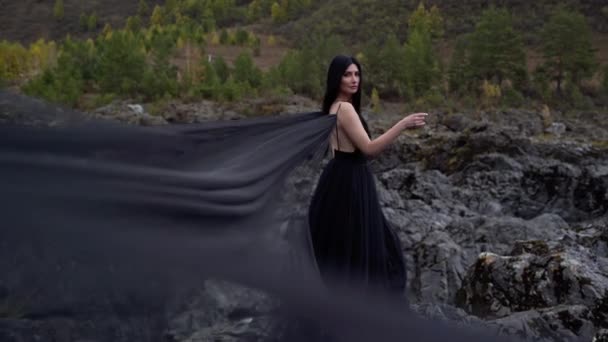 Hermosa chica con el pelo negro se levanta en vestido largo en la montaña del acantilado. La tela se desarrolla en el viento — Vídeos de Stock