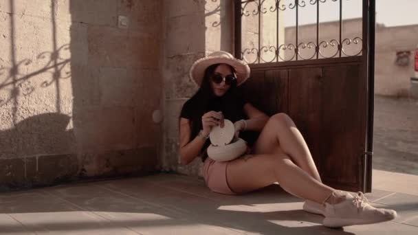 Όμορφη κοπέλα επιρροή σε ένα ψάθινο καπέλο και γυαλιά κάθεται στο έδαφος και προσαρμόζει το μακιγιάζ της. Ζέστη — Αρχείο Βίντεο