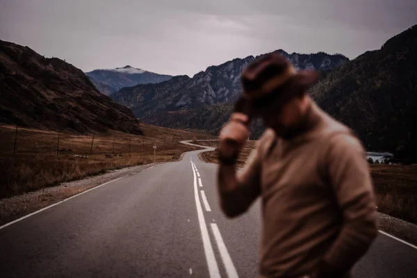 O tipo elegante jovem em um chapéu está no fundo de uma estrada de montanha. Outono Fotografia De Stock