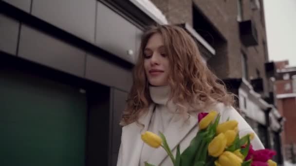 Pěkná holka v bílém plášti s kyticí tulipánů se prochází po ulici. Jaro a tulipány. Úsměv — Stock video