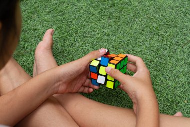 SAMUT-SAKHON, THAILAND - Nisan 04, 2021: Rubik küp bulmacasını çözmeye çalışan genç bir kızın elleri. Çimlerin üzerinde oturuyor..