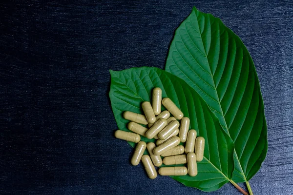 Mitragyna Speciosa Korth veya kratom kapsülleri siyah ahşap masa üzerinde yeşil yaprak, üst görünüm, kopyalama alanı.