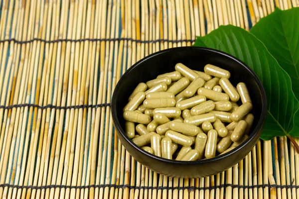 Mitragyna Speciosa Korth or kratom capsules on black bowl with green leaf on wooden mat.