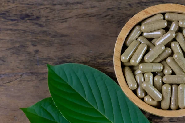 Close-up Mitragyna Speciosa Korth or kratom capsules in wooden bowl with green leaf on rustic wooden table, top view.