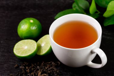 Hot tea in white cup with fresh lime and green leaf on black wooden background.