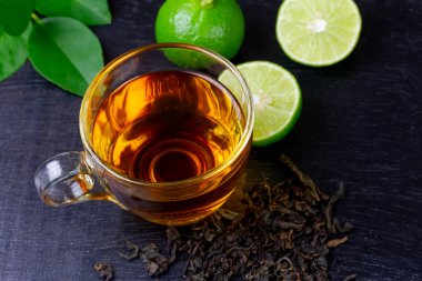 Hot tea in glass cup with fresh lime and leaf on black wooden background.