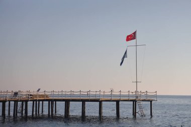 Alanya sahilinde iskele, direkte Türk bayrağı, sakin deniz, açık mavi gökyüzü.