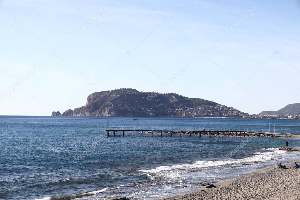 Foam surf on a sandy beach and a pier against the backdrop of a historic mountain in Alanya, November 2021.