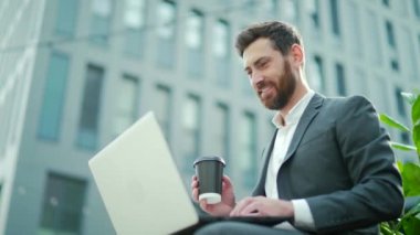 Kafkasyalı sakallı iş adamı laptopla ve açık havada kahve molası üzerinde çalışıyor. Şehir merkezindeki bir bankta oturan bir erkek ofis çalışanı modern bir şehir arka planında, dışarıdaki bir iş yerinde.