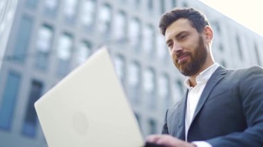 Genç sakallı iş adamı bankta oturuyor ve şehir merkezindeki modern kentsel sokak arka planında dizüstü bilgisayarla çalışıyor.