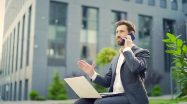 Genç çalışan iş adamı, bankta oturan serbest çalışan, modern kentsel sokak arka planında dizüstü bilgisayarla çalışan şehir merkezindeki girişimci dışında cep telefonuyla konuşan