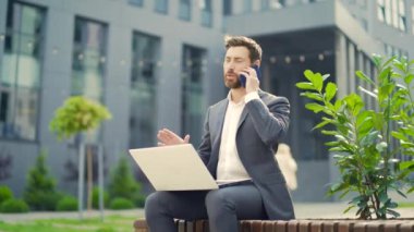 Genç çalışan iş adamı, bankta oturan serbest çalışan, modern kentsel sokak arka planında dizüstü bilgisayarla çalışan şehir merkezindeki girişimci dışında cep telefonuyla konuşan