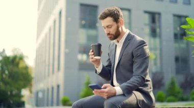 Yakışıklı, mutlu sakallı iş adamı elinde fincan kahveyle bankta oturuyor cep telefonu kullanıyor elinde akıllı telefon, iyi haberler okuyor ve neşeli bir gülümseme. Modern bir şehir arka planının dışında. Şehir parkı