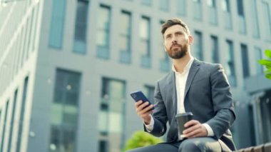 Sakallı yakışıklı iş adamı elinde kahve fincanı, elinde cep telefonu, elinde akıllı telefon ile bankta oturuyor ve akıllı telefon kullanma haberlerini okuyor. Şehir merkezinde. Modern bir şehir arka planının dışında. Şehir parkı