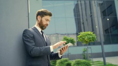 Resmi takım elbiseli şık sakallı iş adamı dışarıda arka plandaki modern ofis binasında elinde tabletle çalışıyor. Adam akıllı telefon kullanıyor ya da şehir dışında cep telefonu kullanıyor.