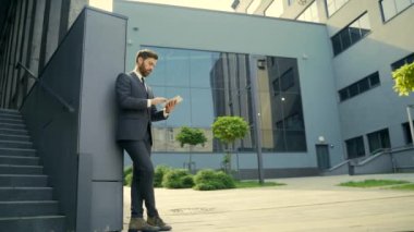 Resmi takım elbiseli şık sakallı iş adamı dışarıda arka plandaki modern ofis binasında elinde tabletle çalışıyor. Adam akıllı telefon kullanıyor ya da şehir dışında cep telefonu kullanıyor.