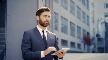 Resmi takım elbiseli şık sakallı iş adamı dışarıda arka plandaki modern ofis binasında elinde tabletle çalışıyor. Adam akıllı telefon kullanıyor ya da şehir dışında cep telefonu kullanıyor.