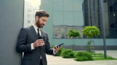 Molada resmi takım elbiseli sakallı iş adamı cep telefonu kullanıyor. Şehir merkezindeki modern şehir sokaklarının arka planında elinde kahve fincanıyla duran bir iş adamı. Boşluğu kopyala
